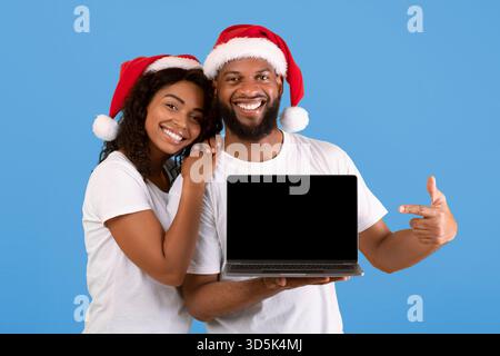 Christmas sale. Excited african american spouses showing laptop with blank screen, posing in Santa hats Stock Photo