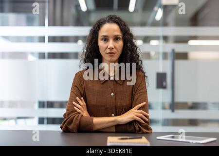 Female professional with dark curly hair and a serious expression sitting at an office desk, arms crossed, confidently looking at the camera during a business meeting or interview Stock Photo