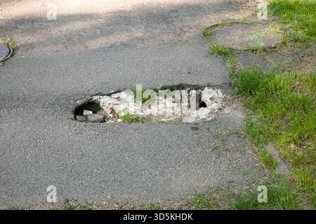 Asphalt with a hole where small green grass grows. Nearby is a grassy lawn. A manhole cover is also visible in the asphalt. Stock Photo