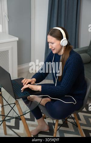 Young woman wearing headphones working from home is having problems with her laptop, she is looking at the screen and typing on the keyboard with a wo Stock Photo