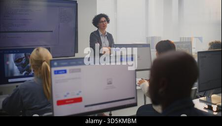 Female Teacher Stands By Large Screen Displaying Programming Code, Explaining Modeling Concepts to Adult Students. Concept Digital Education, Coding Skills, Modern Professional Learning Environment. Stock Photo