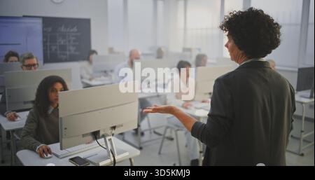 Experienced Female Lecturer Conducts IT Training. Curly Woman Standing and Explaining Task to Adult Students Working on Computers. Concept Technology Education and Knowledge Sharing. Back View. Stock Photo
