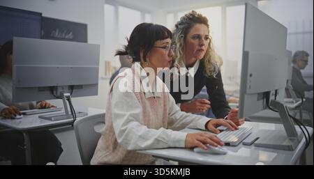 Professional Woman Educator Leads Programming Lesson For Adult Students. Teacher Stands Next Female Student, Checks Progress, Encourages Creative Problem Solving. Concept Modern Education, Motivation. Stock Photo