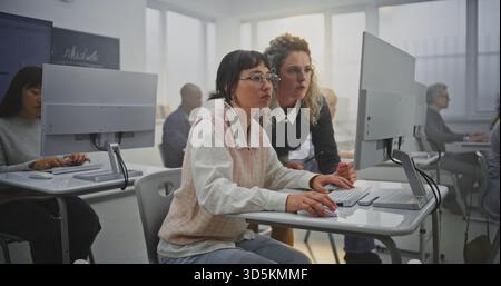 Elegant Female Teacher With Curly Hair Reviews Coding Projects With Adult Students in Bright Modern IT Classroom. Concept Digital Transformation in Education, Support, and Teamwork in Learning. Stock Photo