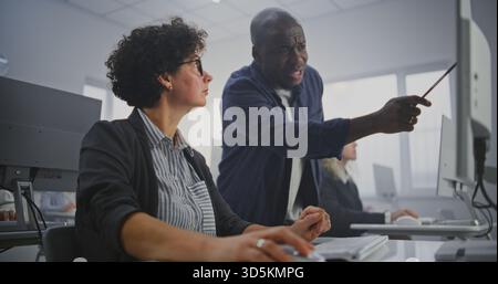 Attentive African American Instructor Walks Between Adult Students During Programming Workshop in Modern Computer Classroom. Helps Debug Code, Provides Feedback, Explains Complex Concepts. Dolly Shot. Stock Photo