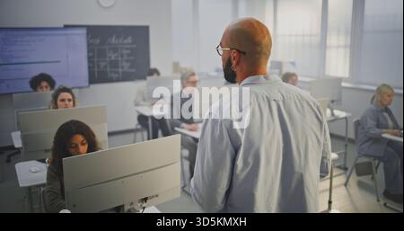 Professional Male Instructor Stands in Front of Adult Students in Computer Lab, Explaining Coding Principles. Concept Continuous Education, Digital Skills, and Modern Learning Environment. Back View. Stock Photo