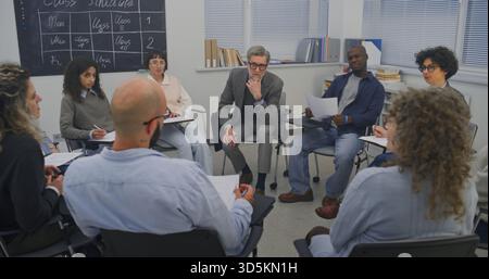 Inside Training Room, Adult Participants Gather in Seated Circle While Instructor Leads Focused Conversation. Students Appear Engaged, Taking Turns Listening and Contributing Dialogue. Dolly Shot. Stock Photo