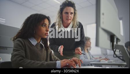 Stylish Mature Female Teacher Walks Through Modern Computer Classroom, Guiding Adult Students During Programming Workshop. Helps Debug Code, Explains Logic, and Fosters Confident Learning Atmosphere. Stock Photo