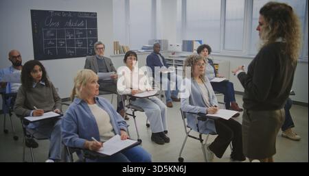 Diverse Adult Learners Sit Modern Desks, Writing and Listening Attentively Teacher. Class Remains Focused, Completing Assignments and Taking Notes During Structured Educational Sessions. Static Shot. Stock Photo