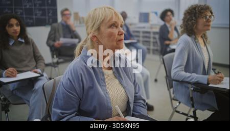 Adult Students Sit Desks, Focusing on Written Tasks While Listening Instructor. Diverse Learners Stay Engaged, Taking Notes and Completing Assignments in Calm, Structured Classroom Environment. Stock Photo
