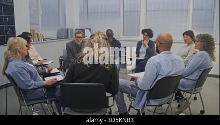 In Classroom With Large Windows and Chalkboard Schedule on Wall, Circle Adult Learners Listens Closely Facilitator. Everyone Seated on Chairs, Participating in Group Exercise or Guided Conversation. Stock Photo