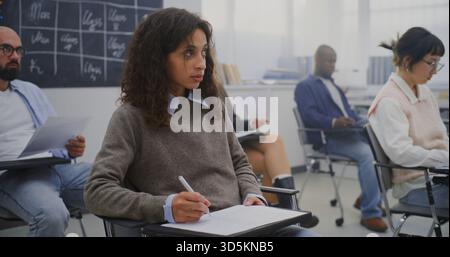 Young Woman in Good Mood Among Group Adult Students of Various Ages Completing Written Test. Students Sitting at Individual Desks and Writing Exam. Concept Professional Training and Lifelong Learning. Stock Photo