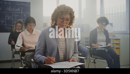 Mature Woman With Curly, Greying Hair Sits at Desk, Deeply Focused on Written Test Among Other Adult Students. Concept of Lifelong Learning and Adult Education in Bright, Modern Setting. Static Shot. Stock Photo