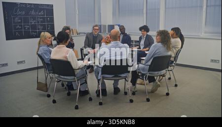 Diverse Group of Adult Students Sits in Circle Inside Modern Classroom. Engaged in Active Discussion With Instructor, Who Gestures While Speaking. Atmosphere Feels Open, Attentive, and Collaborative. Stock Photo