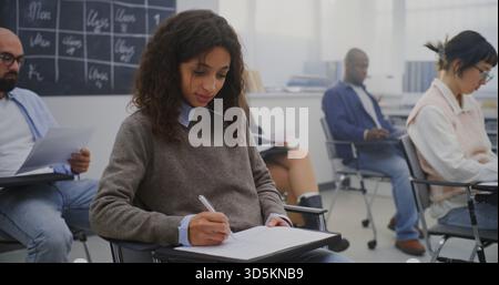 Young Woman in Good Mood Among Group Adult Students of Various Ages Completing Written Test. Students Sitting at Individual Desks and Writing Exam. Concept Professional Training and Lifelong Learning. Stock Photo