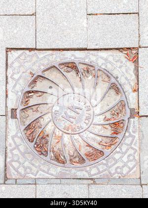 A decorative round manhole cover located on a paved sidewalk is decorated with a pattern resembling diverging petals. Stock Photo