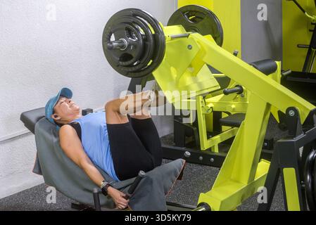 A middle-aged woman in the gym using a leg machine, wearing a cap and blue sportswear. Stock Photo