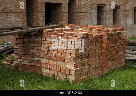Pile of organized red clay bricks stacked outdoors near a building, ready for construction use. The setting suggests ongoing development and preparati Stock Photo