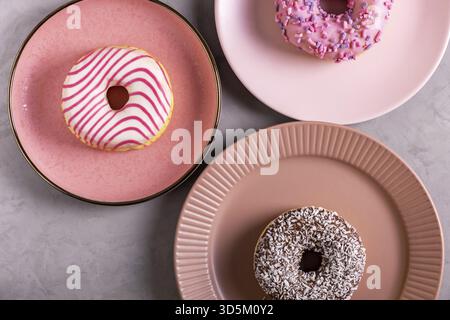 Three glazed donuts lie on tall pink plates on a gray surface. Selective focus. View from above Stock Photo