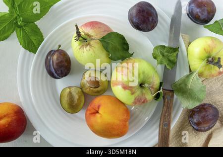 Several ripe fruits and a knife with a wooden handle lie on a white porcelain plate on a light painted wood surface. Horizontal orientation, top view Stock Photo