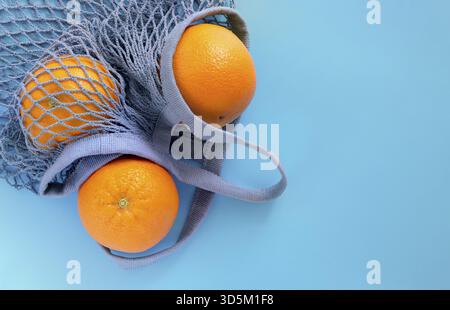 Three ripe oranges lie in a gray-blue eco-friendly string bag on a blue background. Horizontal orientation, selective focus, copy space Stock Photo