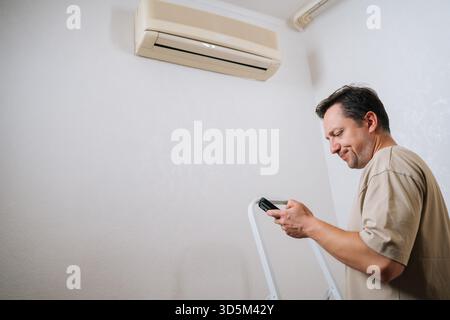 Low-angle view of technician male standing by ladder, using smartphone app to control and prepare for air conditioner maintenance, ensuring optimal Stock Photo