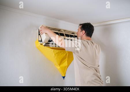 Focused HVAC technician carefully disassembling air conditioning unit, using protective bag to catch dripping water and debris during repair process. Stock Photo