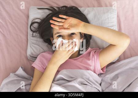 A sick girl lying in bed with a tissue covering her nose, wearing a pink t-shirt, and holding her hand to her forehead, looking unwell. Concept of ill Stock Photo