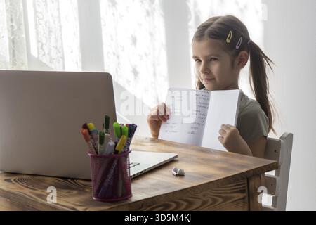 Distance education, schoolgirl studying online on laptop at home, sitting at table and communicating with teacher, shows his notes in his notebook on Stock Photo