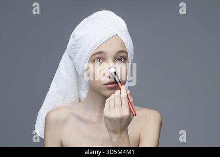 Young caucasian female applying makeup with brush on her face in skincare routine Stock Photo
