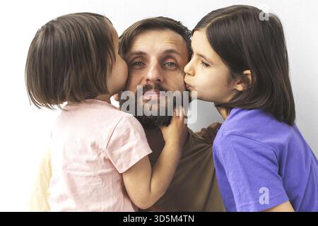 Joyful father smiling while receiving kisses on cheeks from two loving daughters, celebrating family affection and Father's Day against white backgrou Stock Photo