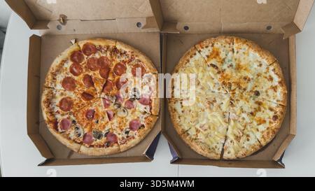 Two whole pizzas in open cardboard boxes on white table, one with pepperoni and mushrooms, the other with pineapple and cheese, concept of delivery an Stock Photo