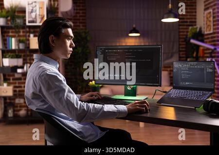 Focused asian man working late, using desktop pc and laptop to debug software. Dedicated remote developer analyzing website code on multiple screens and completing programming tasks at home Stock Photo