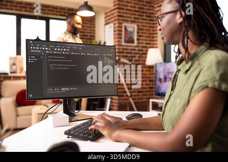 Female programmer sitting at home workstation, focused on writing website code as boyfriend watches television. African american tech entrepreneur developing software remotely on computer. Stock Photo