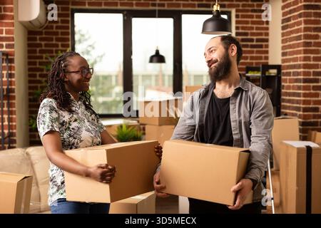 Black woman and white man moving into new home, reflecting exciting beginnings together. Happy multiethnic couple relocates with packed belongings, holding boxes in modern brick wall apartment. Stock Photo