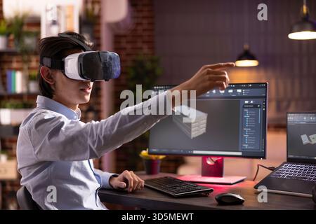 Asian man sits at desk with computer and laptop, using virtual reality to interact with 3d container model in cad software. Young male freelancer with vr headset working on client projects remotely. Stock Photo