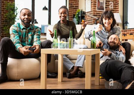 Bearded man and white woman team up with african american friends for online gaming. Multiethnic group reacting joyfully, holding controllers and enjoying fun video game in cozy apartment. Stock Photo