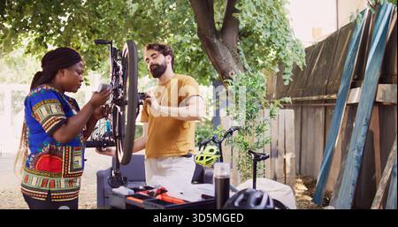 Healthy determined multiracial pair in yard fixing damaged bike components with professional toolkit. Sporty black woman grabbing tool and lubricant for bicycle wheel maintenance with caucasian man. Stock Photo