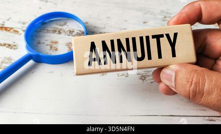 Hand Holding a Wooden Block Engraved with "Annuity" Next to a Magnifying Glass, Representing Retirement Savings and Financial Planning Stock Photo