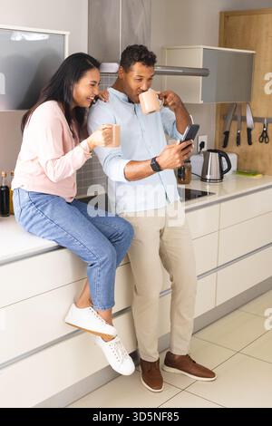 Couple in kitchen drinking coffee and using smartphone, enjoying morning together Stock Photo