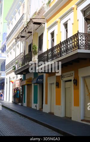 Colorful buildings line a narrow cobblestone street, with ornate wrought iron balconies and vibrant facades in shades of yellow and white. Stock Photo