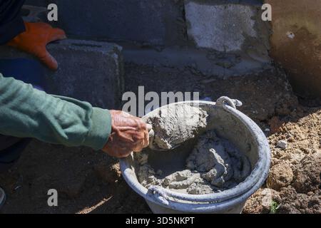 Focused manual labor on construction site. person wearing glove mixes gray cement mortar in bucket for foundation repair, showing hard work and skille Stock Photo