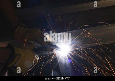 Focused welder at work using welding torch on metal beam. Bright hot sparks fly from industrial process in dark workshop, showcasing skilled manual la Stock Photo