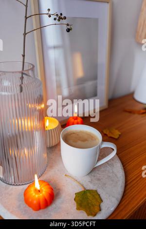 Autumn-inspired setting with a white coffee cup on a stone tray, surrounded by small pumpkins, candles, and a vase, evoking a cozy and inviting ambian Stock Photo