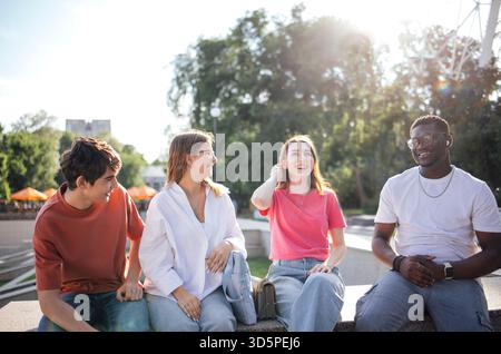 Diverse group of young adults is sitting together outdoors, laughing and sharing stories in a lively urban environment, highlighting joy and camarader Stock Photo