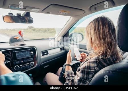 A young Caucasian woman with long blonde hair sits in a car, using her smartphone. A man drives, wearing a blue cap. The scene captures a journey on Stock Photo