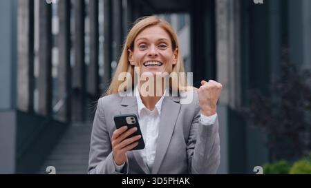 Surprised happy Caucasian adult 40s business woman with phone win in city amazed winner businesswoman winning with mobile app yes gesture female Stock Photo