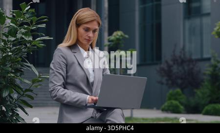 Focused Caucasian middle-aged business woman typing laptop in city adult busy businesswoman female working computer online corporate service outdoors Stock Photo