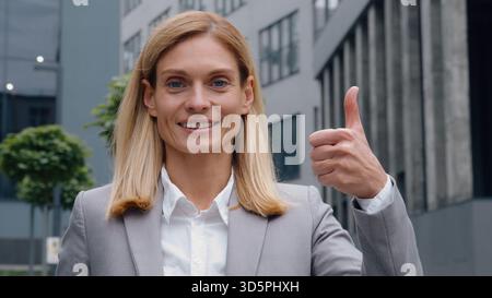 Caucasian middle-aged smiling business woman showing thumb up businesswoman like gesture in city near office building looking at camera employer Stock Photo