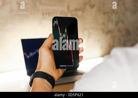 Close-up of a person's hand gripping a smartphone showing a red declining stock market chart, laptop with similar graph visible behind on a desk. Stock Photo